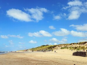 Ein Sandstrand mit blauem Himmel, einem Leuchtturm und bewachsenen Dünen im Hintergrund.