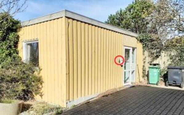 A small, single-story building with a door and window, surrounded by vegetation.
