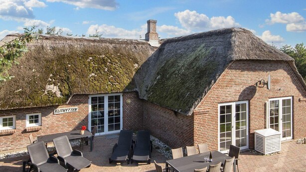 A thatched-roof building with a brick exterior, a patio with lounge chairs, and a table with a bottle. - 4 Star Holiday Home in Ulfborg (Ulfborg)