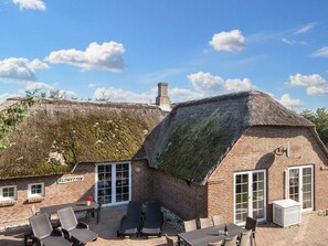 Un bâtiment à toit de chaume, avec une façade en briques, une terrasse dotée de chaises longues et une table sur laquelle se trouve une bouteille.