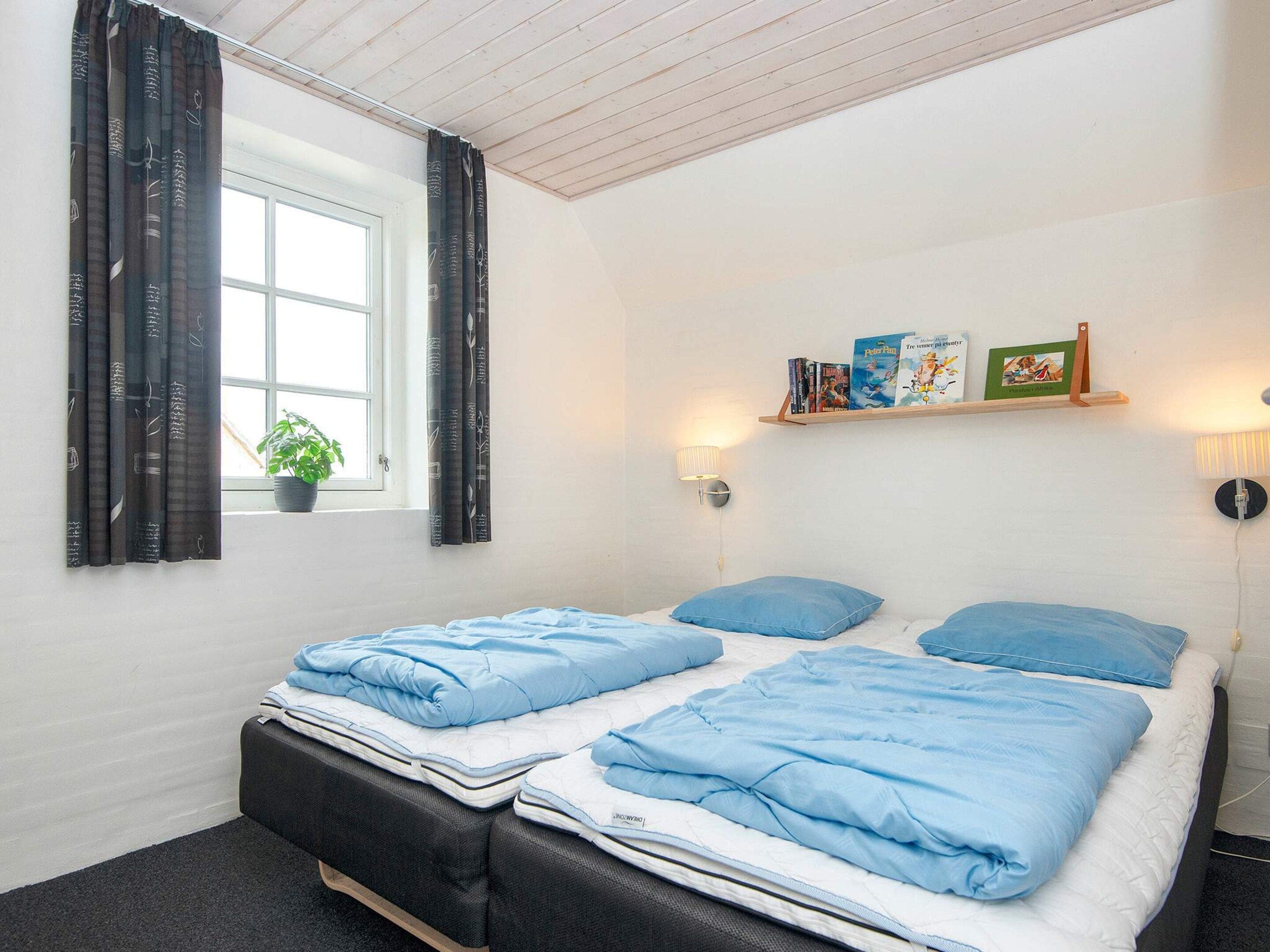 A bedroom with a bed, a window with curtains, a bedside table with a lamp, and a shelf with books.