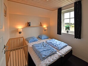 Un dormitorio con una cama doble, dos lámparas de noche, una cuna de madera, una ventana con vistas a la vegetación y una estantería con libros y una lámpara.