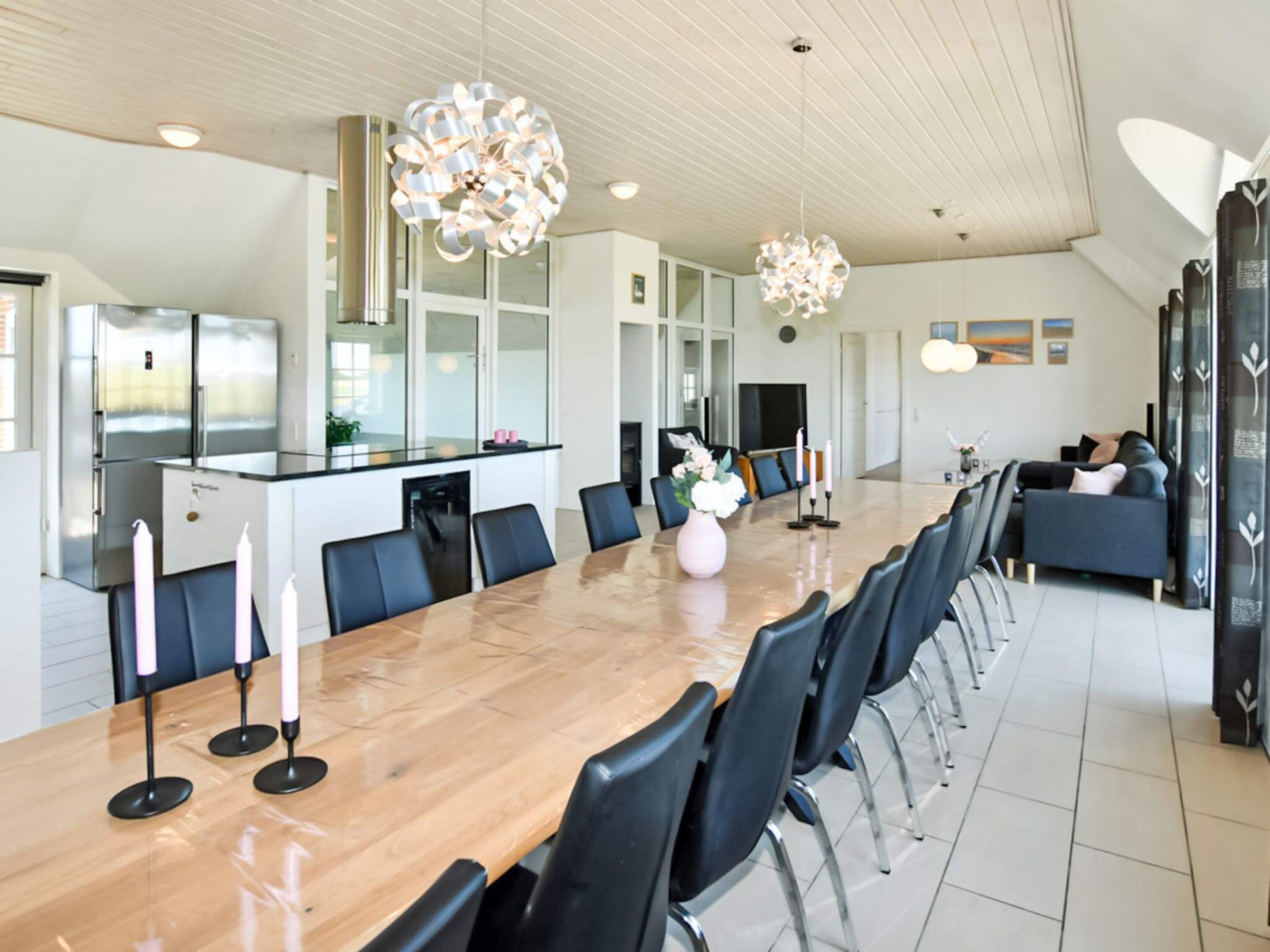 A modern kitchen with stainless steel appliances, a dining area with a long wooden table and black chairs, and a living area with a sofa and decorative pillows.