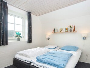 Un dormitorio con una cama, una ventana, una estantería con libros y objetos decorativos, y una lámpara fijada a la pared.