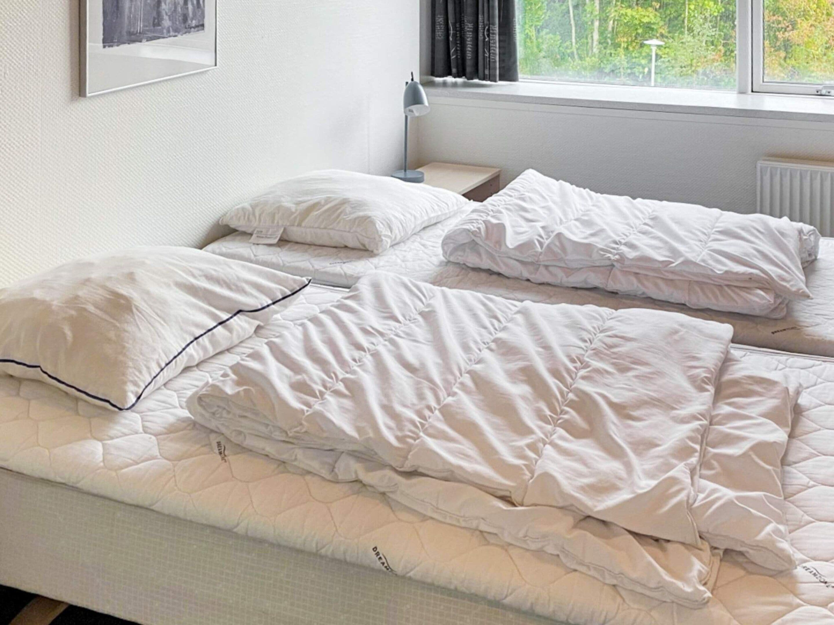Una cama individual con colchón acolchado y almohadas, una mesita de noche con lámpara, y una ventana con cortinas.