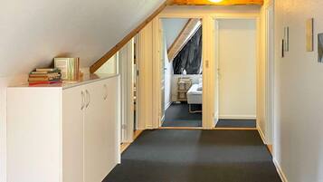Un estrecho pasillo con piso alfombrado, vigas de madera en el techo y una puerta que da a un dormitorio con cama y ventana.