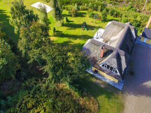 Une maison à toit de chaume, dotée d’un toit en pente, entourée d’un jardin bien entretenu où poussent différentes espèces de plantes et où se trouve une balançoire.