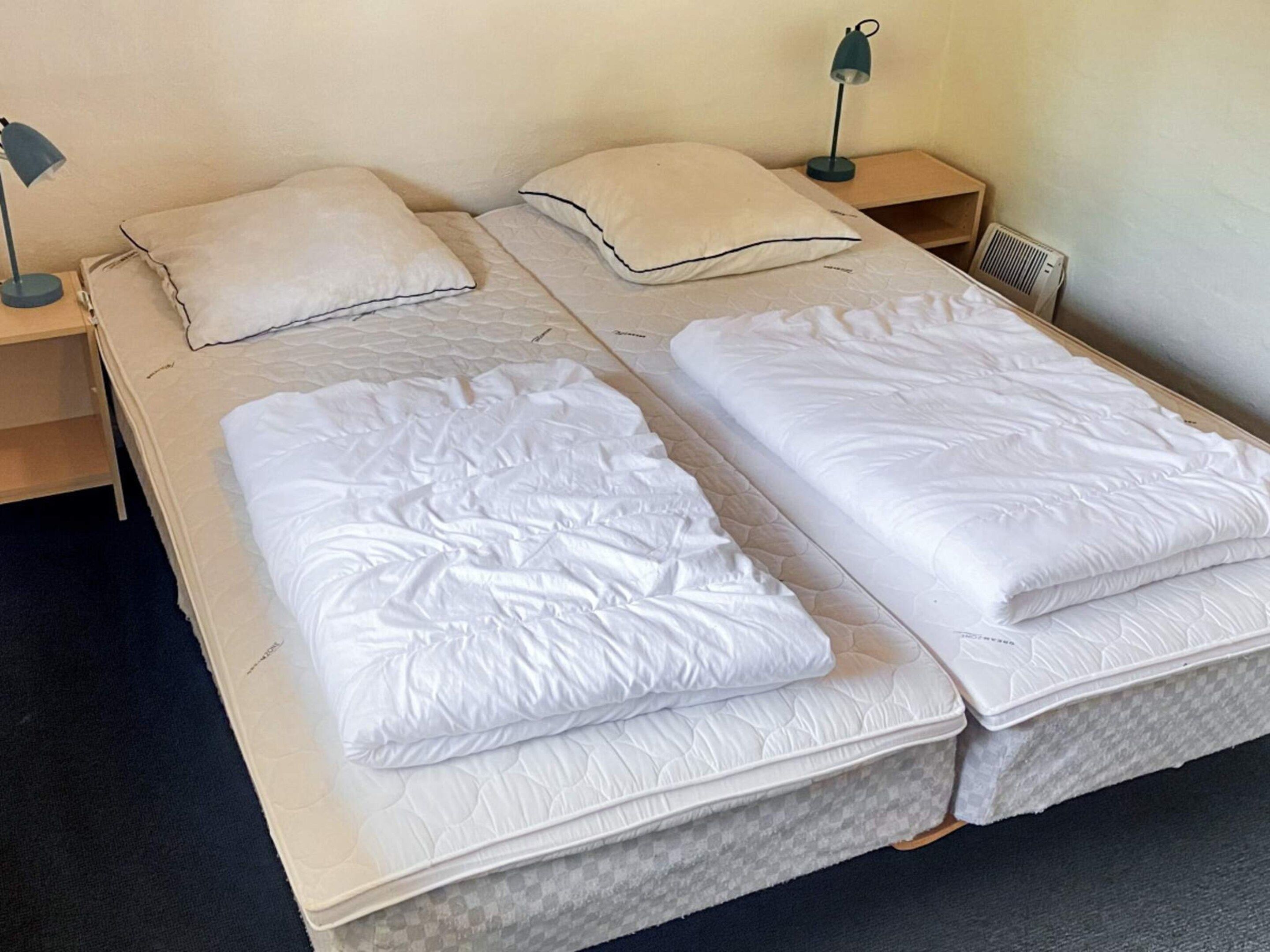Een tweepersoonsbed met een witte dekbedovertrek en kussens, een nachtkastje met een lamp, en een luchtventilation aan de muur.