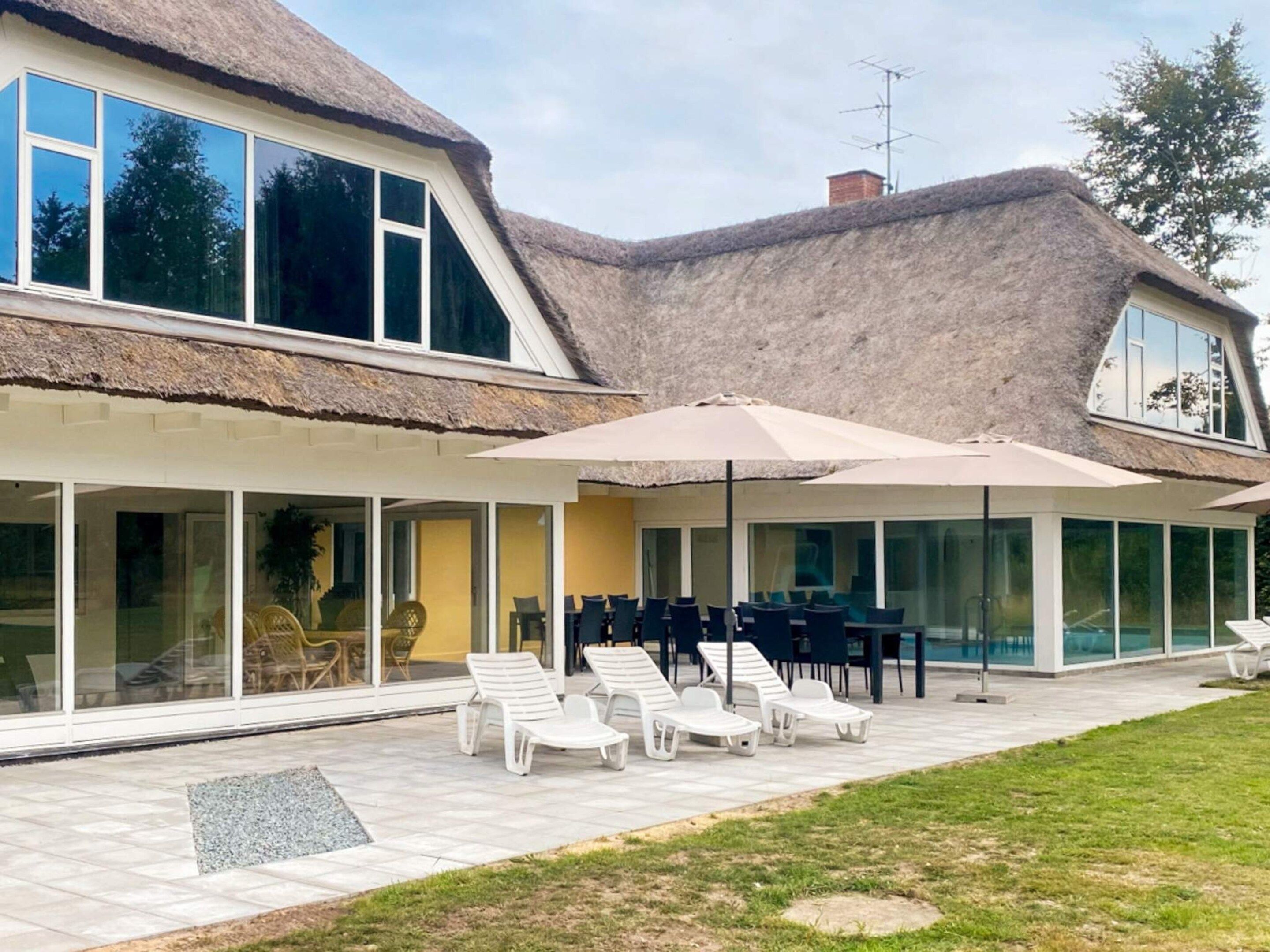 Une maison moderne avec un toit de chaume, de grandes fenêtres et un espace extérieur aménagé avec des fauteuils et des parasols.