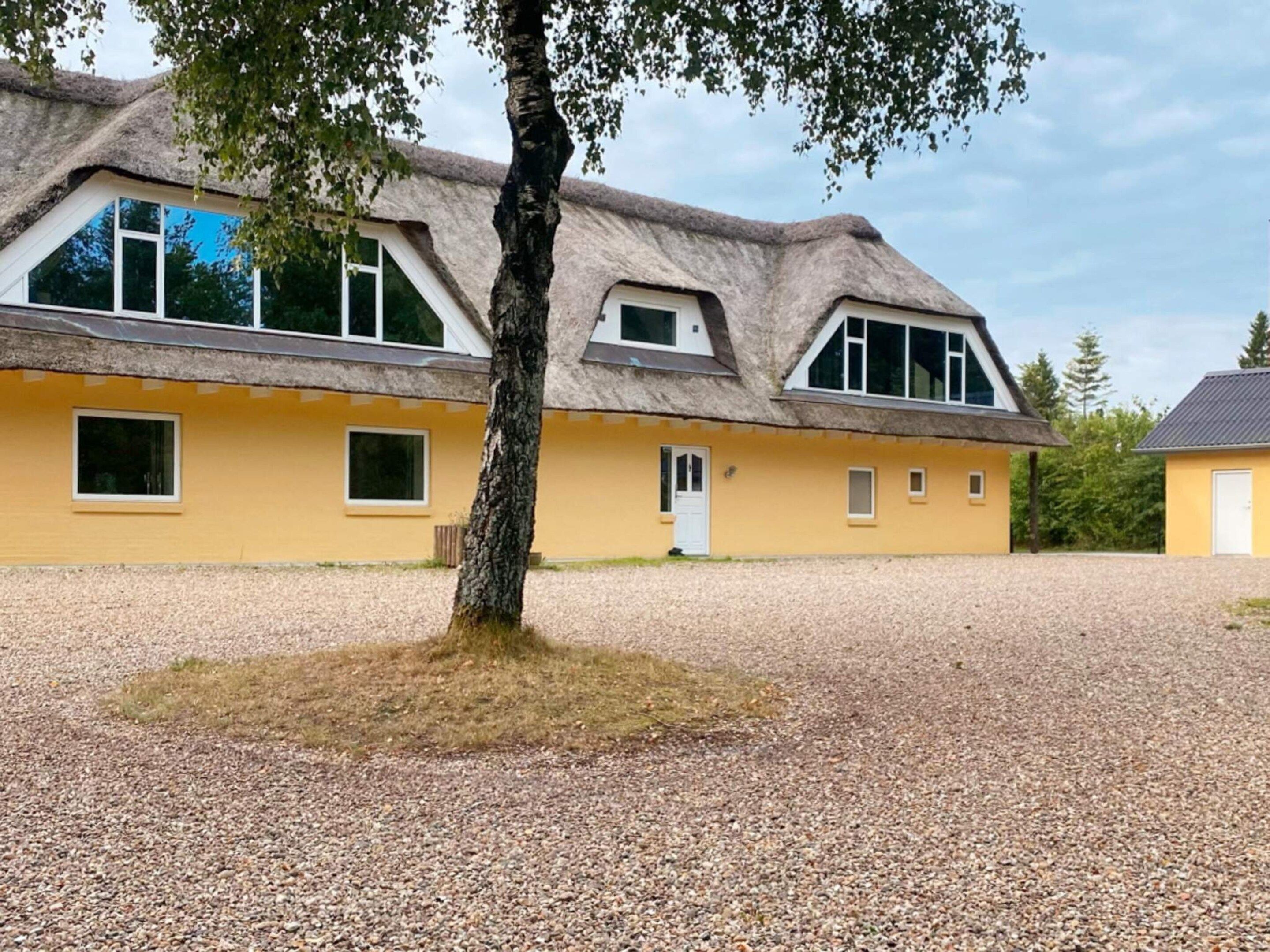 Un bâtiment jaune avec un toit de chaume, une allée de gravier et un arbre au premier plan.