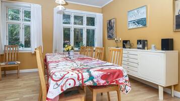 A dining room with a table set for four, flanked by two windows with curtains, a white cabinet with a radio and a candle, and a painting on the wall.