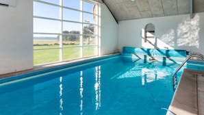 Piscina interna com janelas de vidro transparente, ventilador de teto e vista para uma paisagem verde.