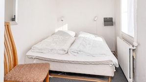 Uma cama de solteiro com edredom branco, uma cadeira de madeira, uma mesinha e um radiador.