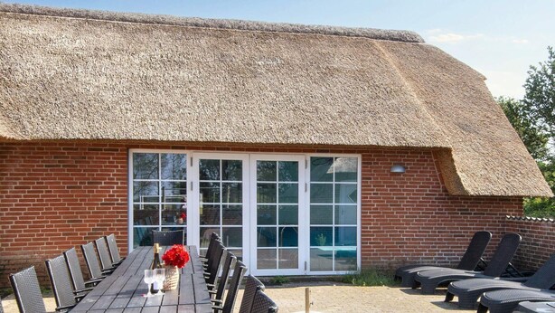 A thatched-roof building with a brick exterior, a large glass door, and a table with chairs and a bottle of champagne. - Luxury Pool House by Sea - By Traum Ferienwohnungen (Ulfborg)