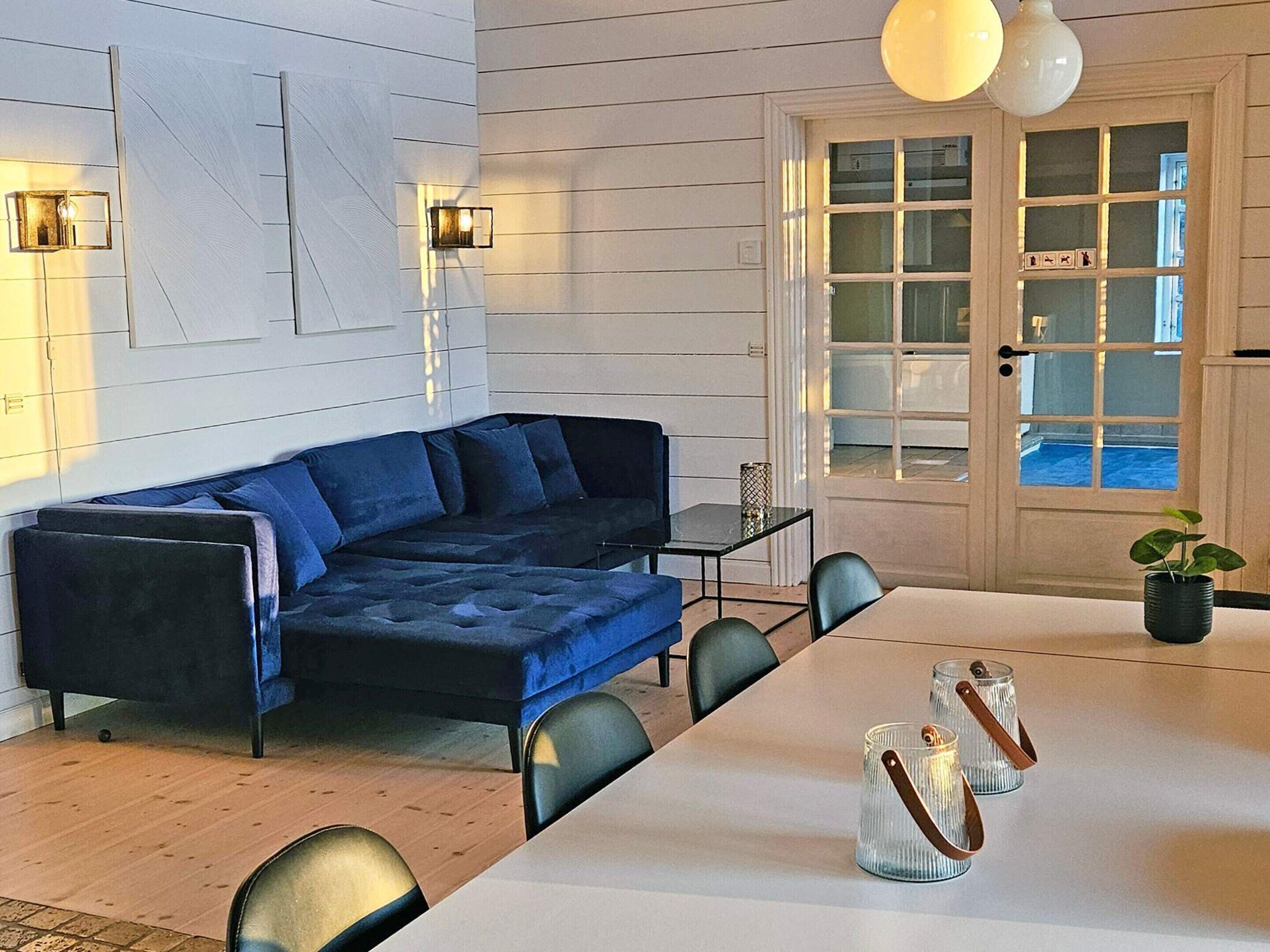 A modern living room with a blue sectional sofa, a dining table with chairs, and a large window with a view of a pool.
