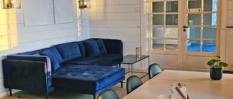 Een moderne woonkamer met een blauwe hoekbank, een eettafel met stoelen en een groot raam met uitzicht op een zwembad.