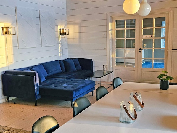 A modern living room with a blue sectional sofa, a dining table with chairs, and a large window with a view of a pool.