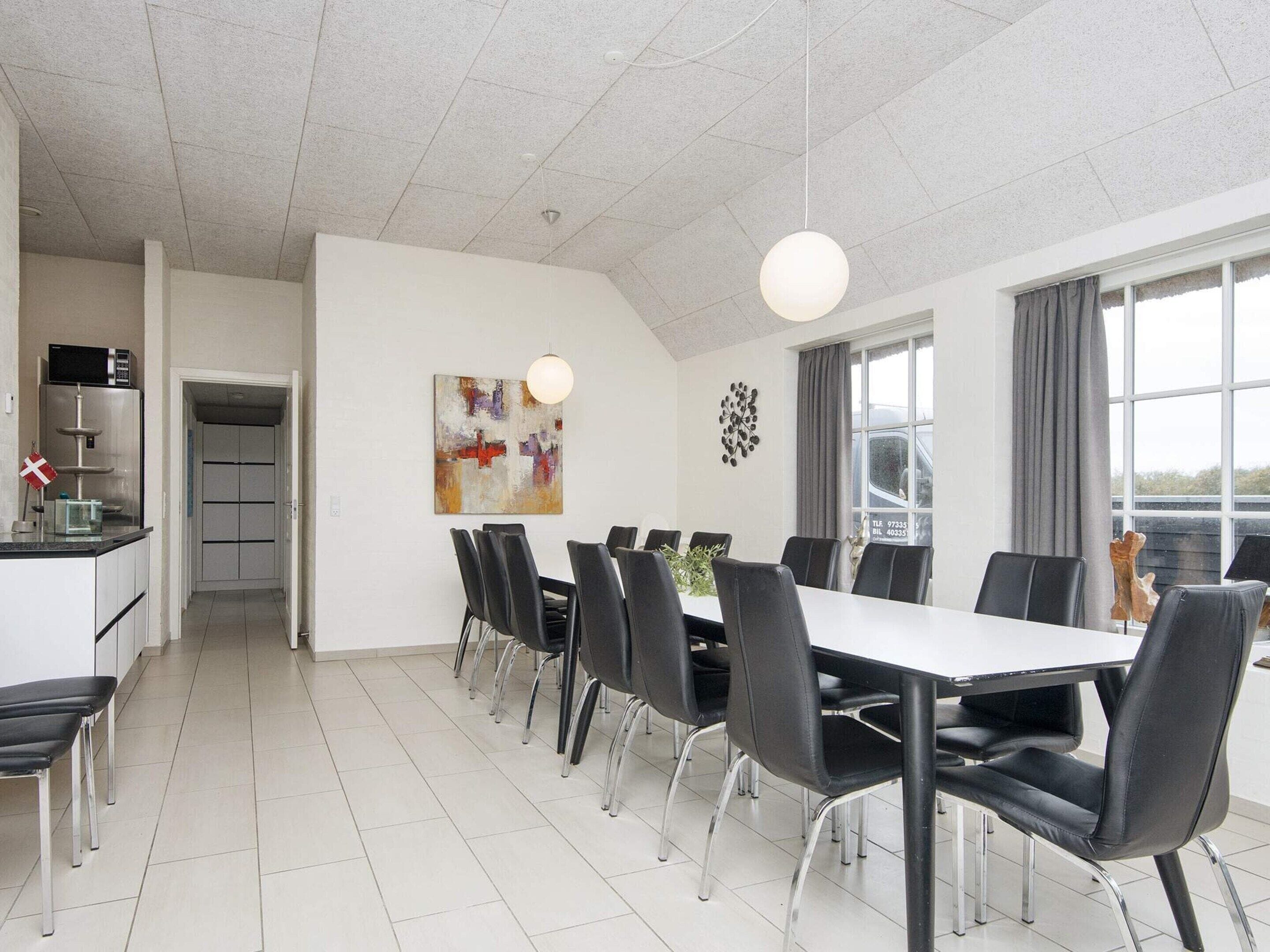 A modern dining area with a long table and chairs, a kitchen area with appliances, and a large window providing natural light.