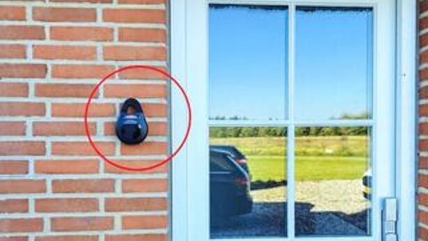 A black security camera mounted on a brick wall, a glass door with a view of a parking lot, and a clear blue sky. - 4 Star Holiday Home in Ulfborg (Ulfborg)