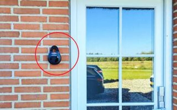 A black security camera mounted on a brick wall, a glass door with a view of a parking lot, and a clear blue sky. - 4 Star Holiday Home in Ulfborg (Ulfborg)