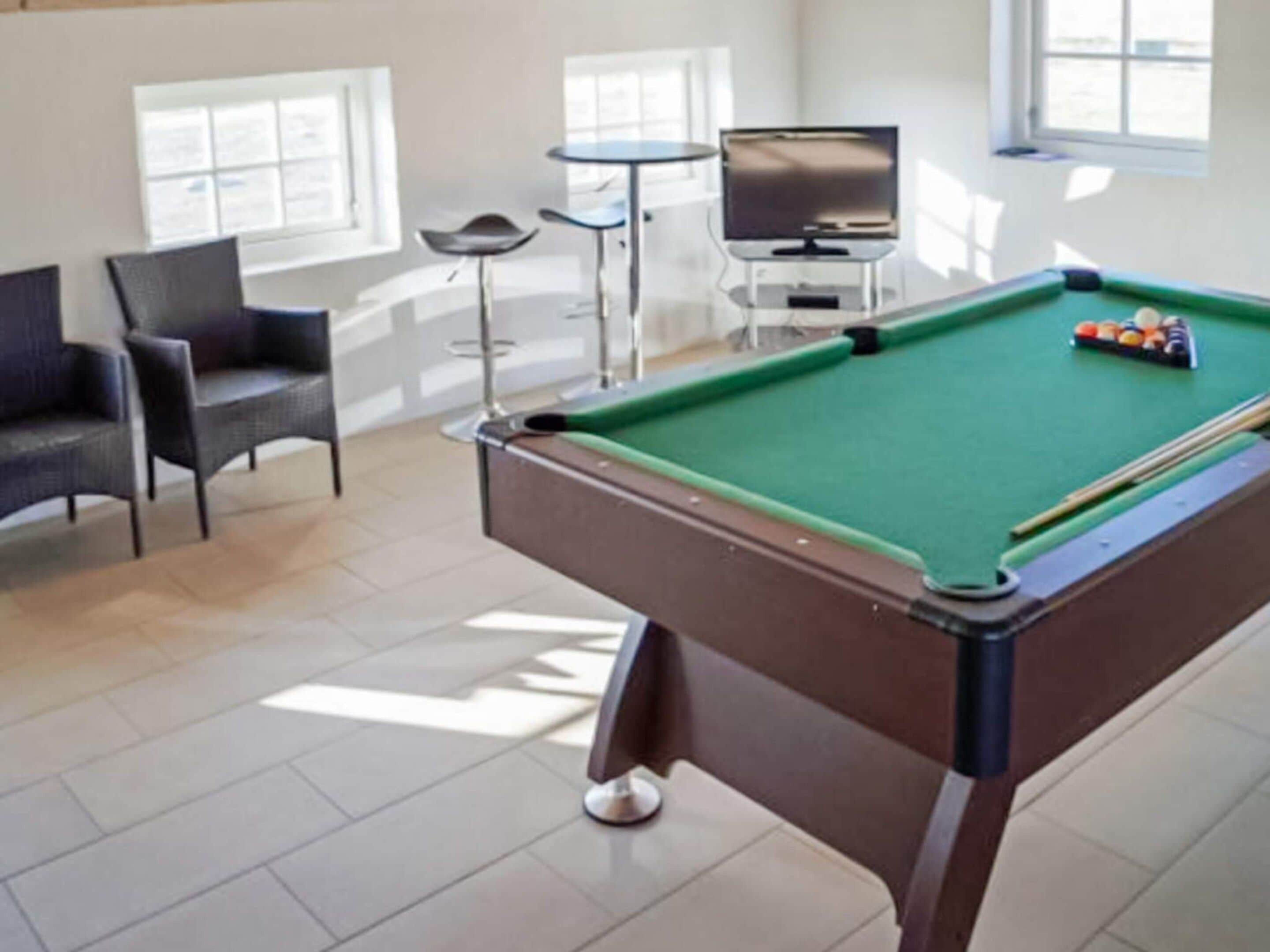 A pool table with a green felt surface, a pool cue, and billiard balls. There are two chairs and a small table with a television in the background.