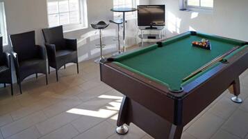 Una mesa de billar con superficie de fieltro verde, rodeada de sillas y un televisor.