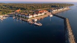 An aerial view of a coastal town with a marina, buildings with red roofs, and a forested coastline.