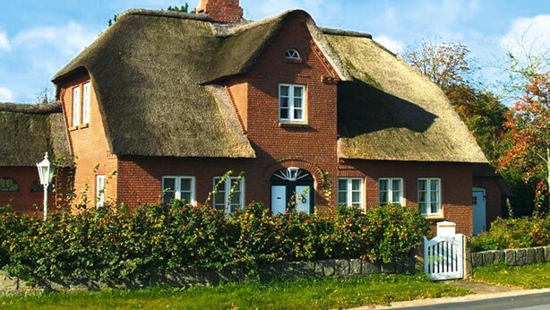 Une maison traditionnelle en briques rouges, avec un toit de chaume et un petit jardin à l’avant.
