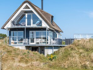 Une maison côtière moderne avec un toit de chaume, de grandes fenêtres et un balcon doté d’un espace salon extérieur.