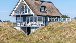 A thatched-roof house with a balcony and large windows, situated on a grassy hill. - 6 Person Holiday Home in Fano (Fano)