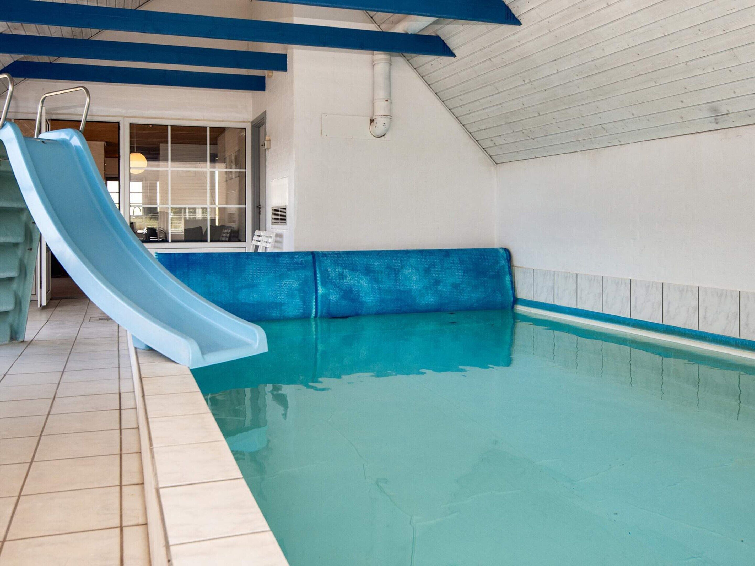 An indoor swimming pool with a blue slide, a tiled floor, and a ceiling with blue beams.