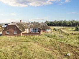 Una casa con techo de paja sobre base de ladrillo, rodeada de campos cubiertos de césped y árboles.