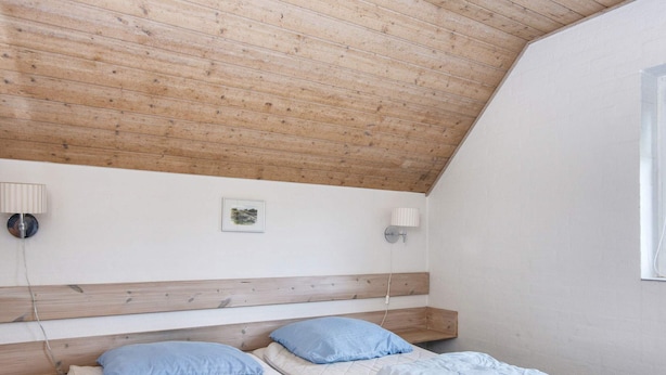 A room with a sloped ceiling, two beds with blue bedding, a wooden headboard, a wall-mounted lamp, and a small framed picture.