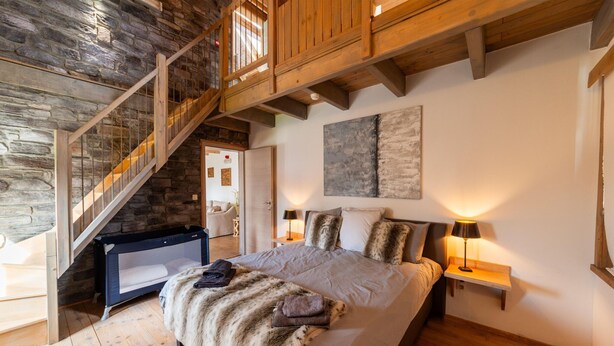 Une chambre avec un mur en pierre, un escalier en bois, un grand lit recouvert de plaids en fourrure et un plancher en bois.