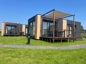 Une maison moderne à deux étages, dotée de grandes fenêtres en verre, d’une terrasse avec des meubles d’extérieur et d’une pelouse.