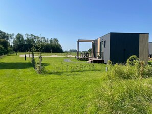 Un bâtiment moderne doté d’une terrasse et d’un espace extérieur aménagé pour s’asseoir, entouré d’un champ verdoyant et d’arbres.