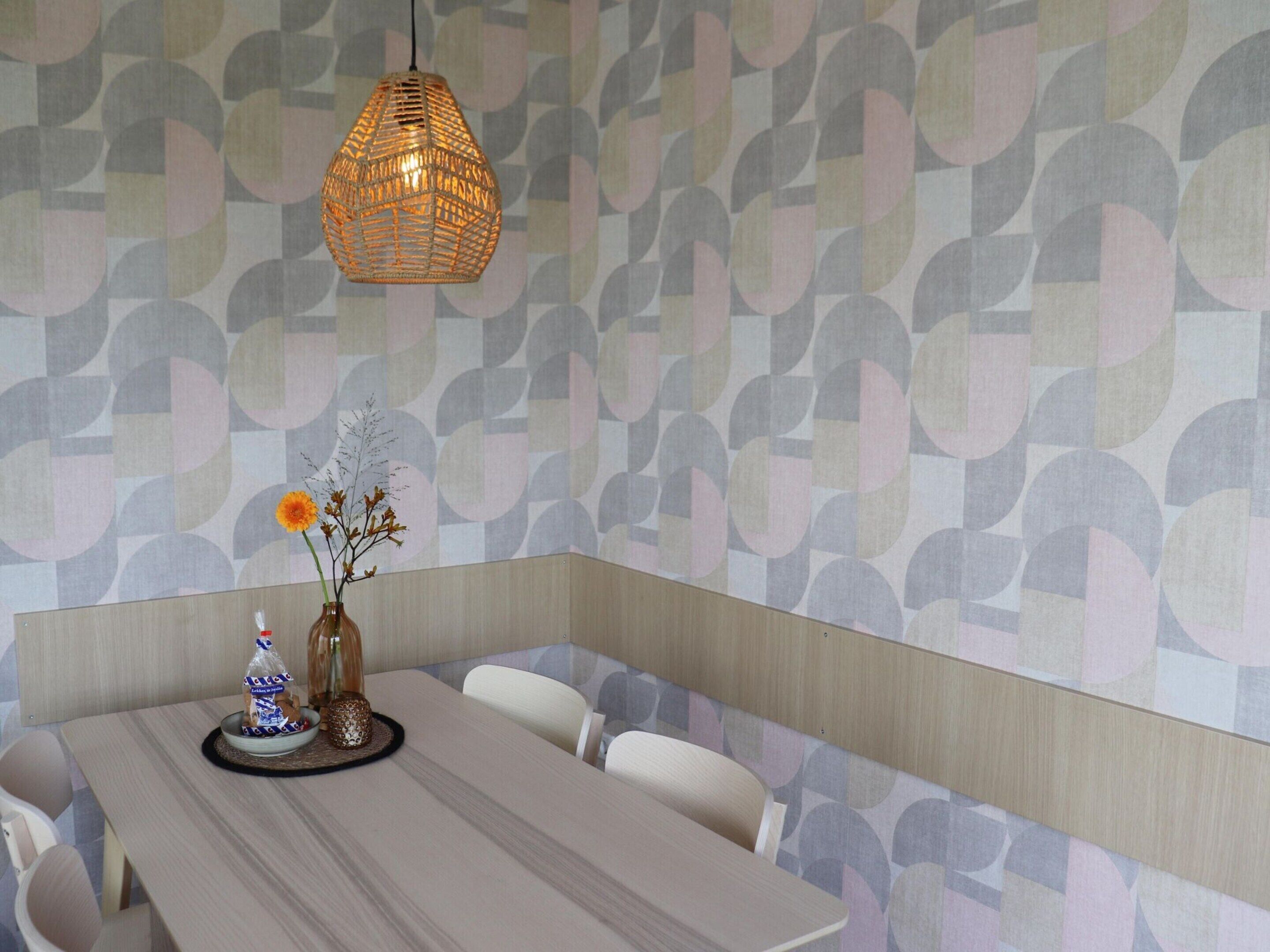 Un espace repas comprenant une longue table, des chaises, un luminaire suspendu et un mur orné d’un motif géométrique.