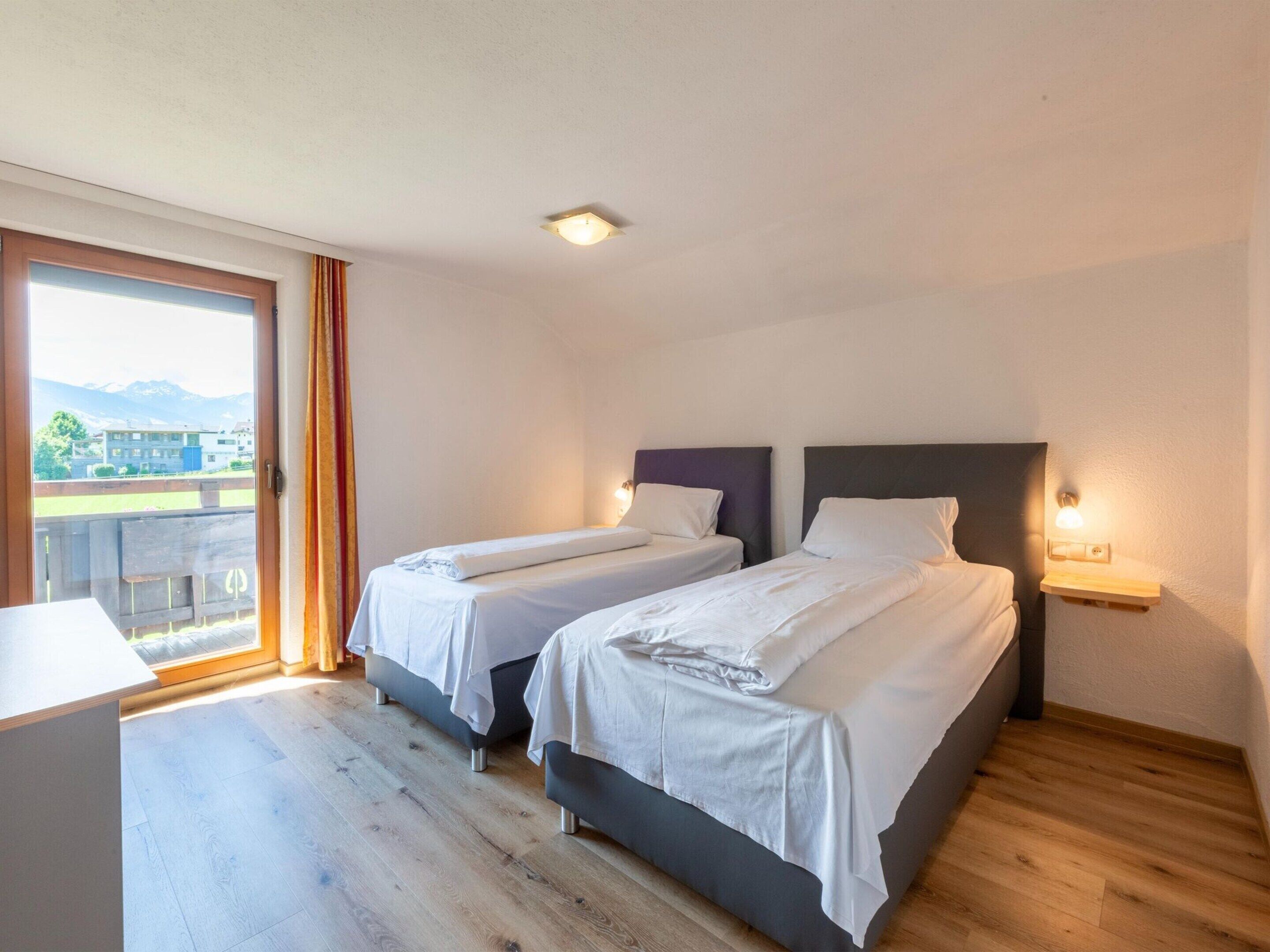 A hotel room with two single beds, a wooden floor, a large window with a view of mountains, and a small desk.