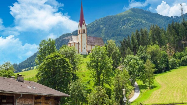 Une église dotée d’un clocher imposant, entourée de collines verdoyantes et d’arbres luxuriants.