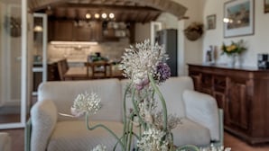 A living room with a sofa, a table with a floral arrangement, and a kitchen in the background. - Belvilla by OYO Casale Degli Ulivi (Lucignano)