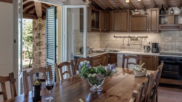 A rustic kitchen with wooden cabinets, a stone wall, and a dining area with a wooden table and chairs. - Belvilla by OYO Casale Degli Ulivi (Lucignano)