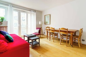 A room with a red sofa, a wooden dining table with chairs, a small side table, and a lamp. - PARI-PEC534 Summary: (Paris)