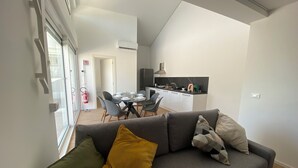 A modern living room with a grey sofa, a dining area with a table and chairs, and a kitchen with white cabinets and a black countertop.