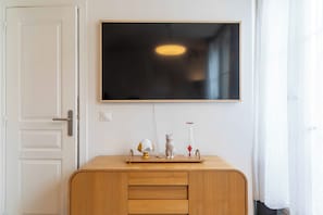 A room with a white door, a wooden cabinet, a flat-screen TV mounted on the wall, and a small table with decorative items. - Beautiful 1BD Apartment in Saint Lazare (Paris)