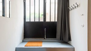 Eine enge Treppe führt zu einer dunklen Tür mit einem Fenster darüber. Am Fuß der Treppe liegt eine kleine, orangefarbene Matte und rechts daneben eine Holzkiste.