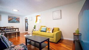 Un salón con un sofá amarillo, una mesa de comedor con sillas, una mesa baja y un mueble para televisión.