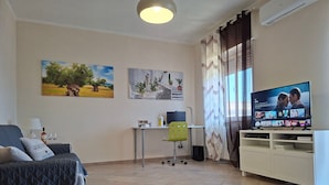 A living room with a grey couch, a TV on a stand, a small table with a lamp, and two paintings on the wall. - The House OF THE SUN Brindisi (Brindisi)