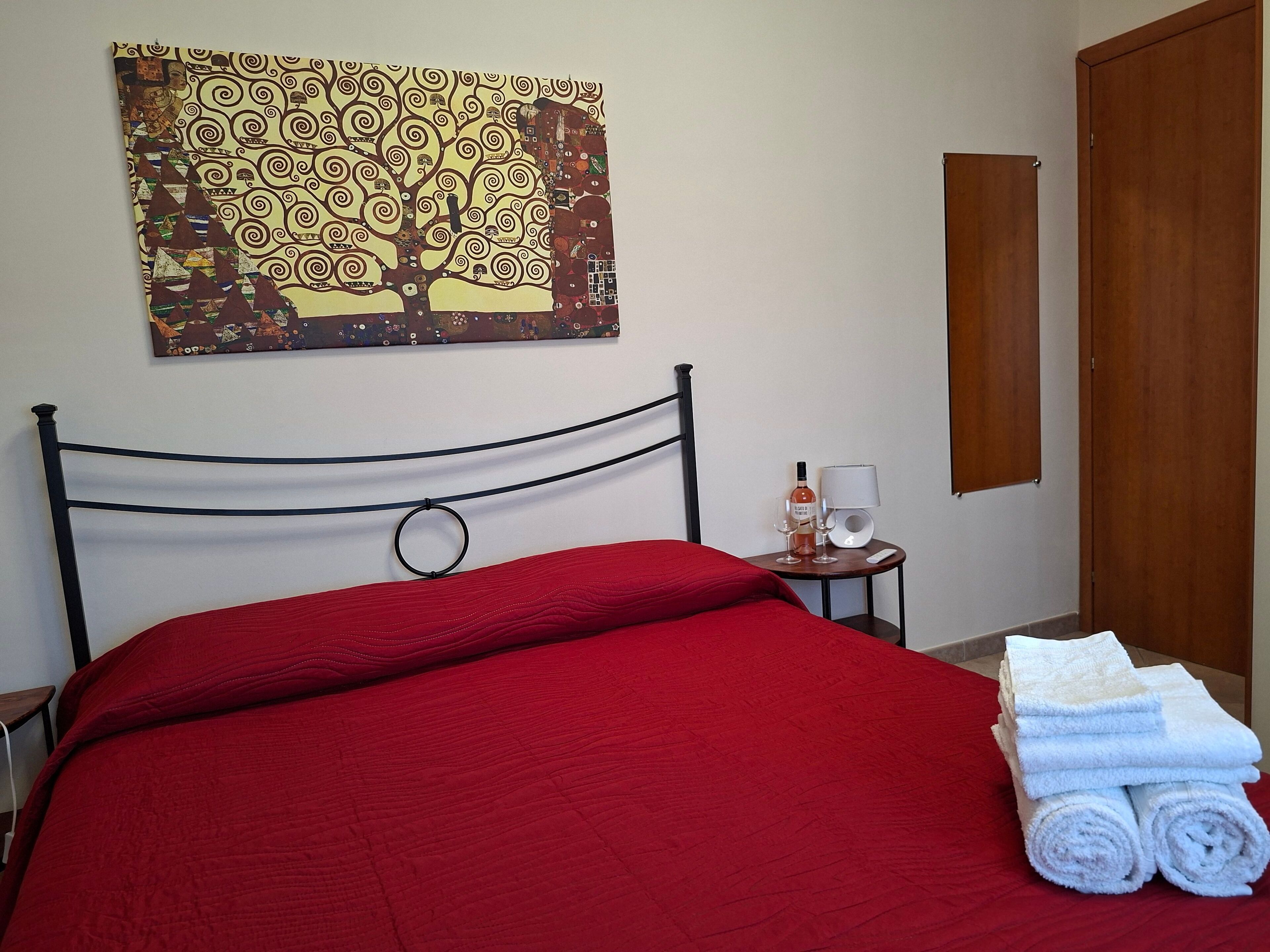 Quarto de hotel com colcha vermelha, um quadro pendurado na parede com uma árvore, mesa de cabeceira com uma garrafa e copos, e uma pilha de toalhas brancas.