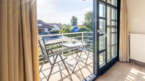 Un balcón con mesa y sillas, vista al vecindario y una ventana con cortinas.