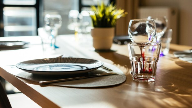 Una mesa de comedor con platos, cubiertos y vasos, en una habitación bien iluminada con una ventana y una planta al fondo.